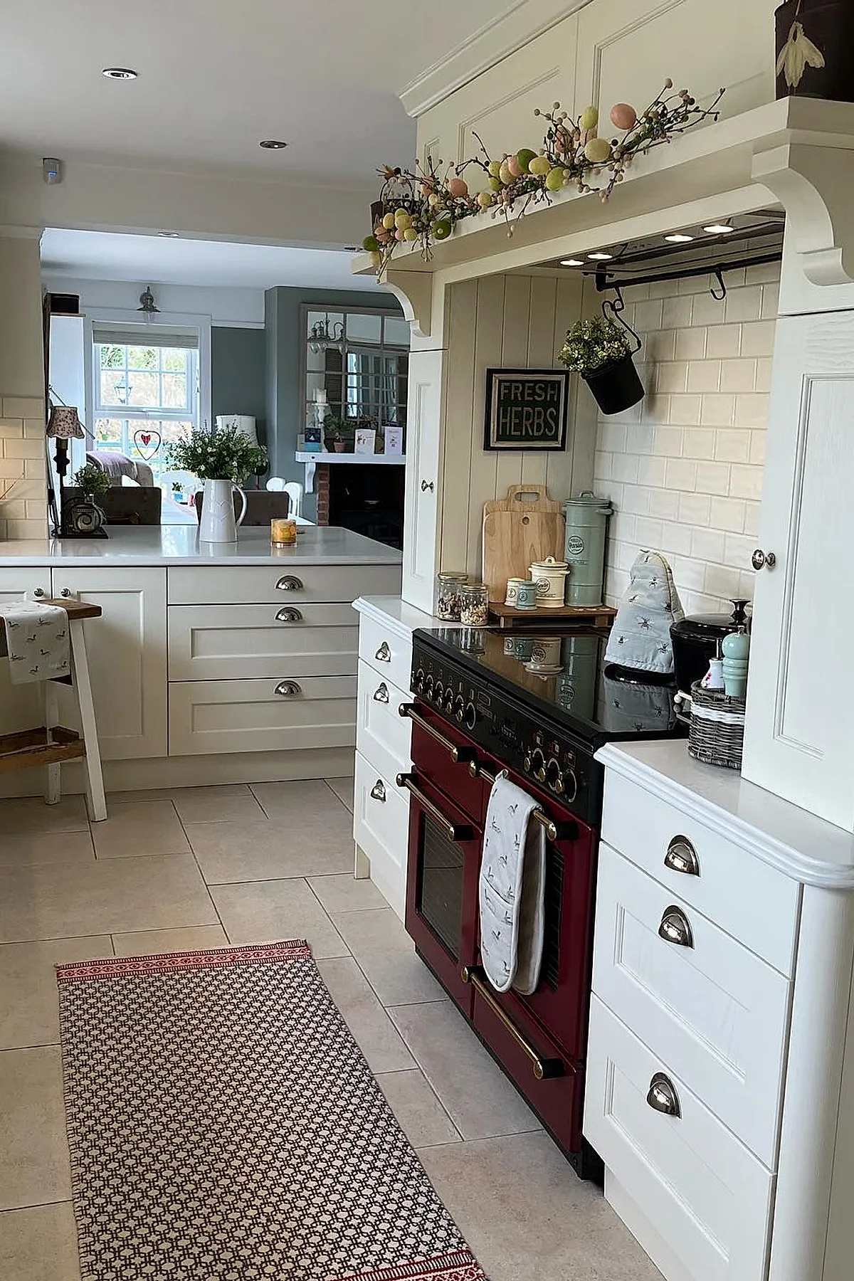 Vintage cottage kitchen featuring a deep red range oven set within white cabinetry, cream tiled floor, white subway tile backsplash, light beige countertop, and decorative floral garland above the stove area.