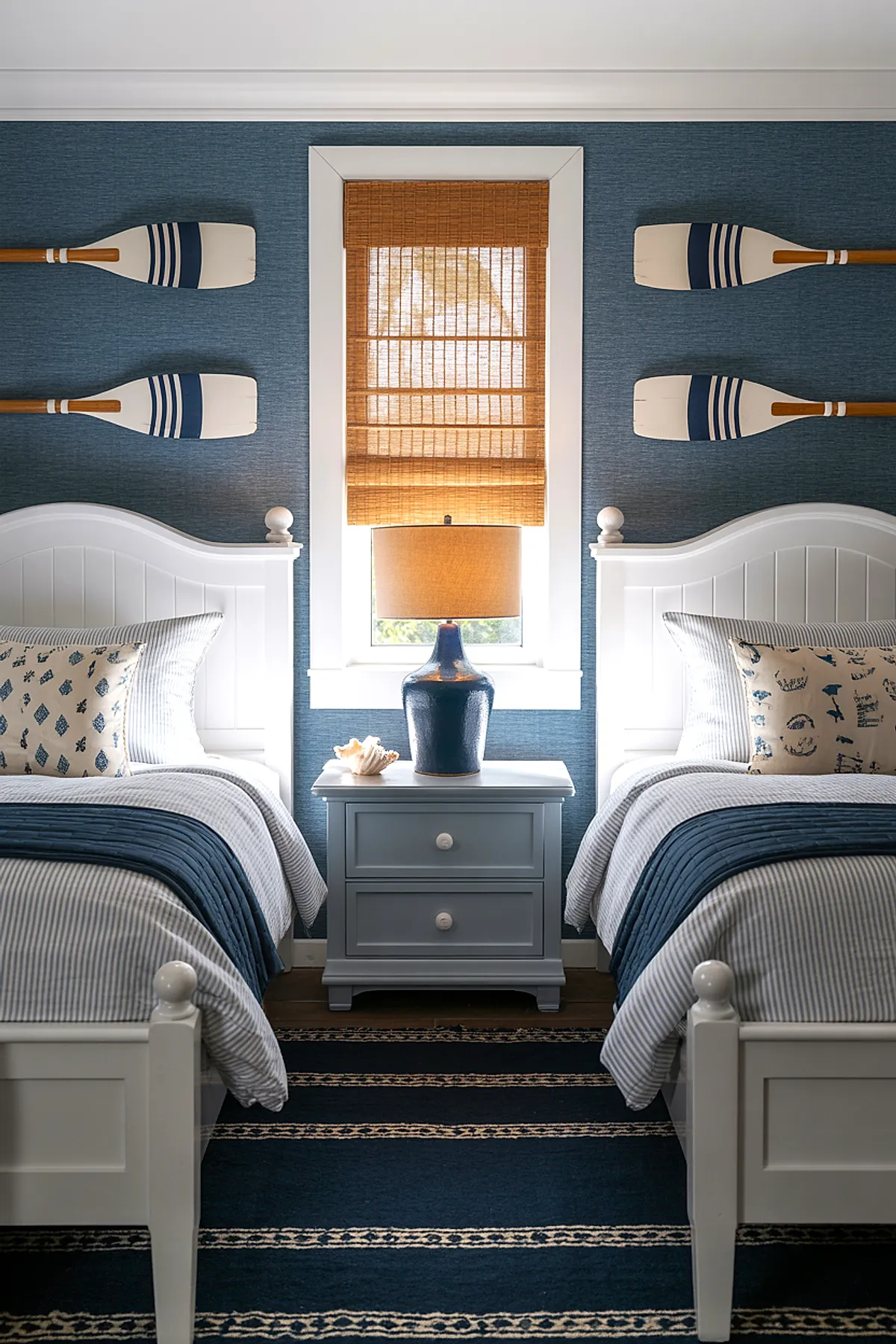 Boys bedroom featuring two white beadboard twin beds with blue and gray striped bedding, a light blue nightstand between them holding a navy ceramic lamp with tan shade, window with bamboo woven shade in center, blue textured wallpaper, and four decorative wooden oars mounted horizontally on walls.