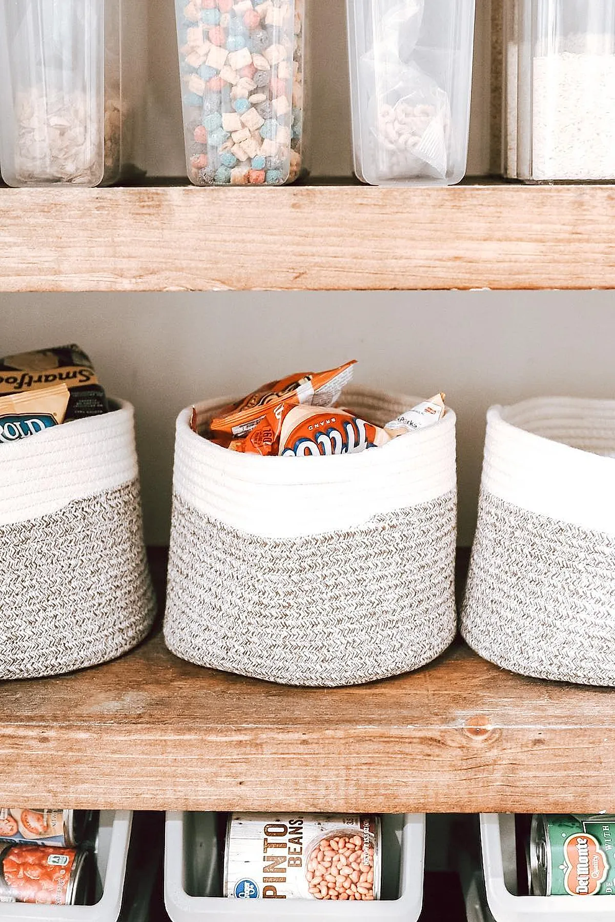 Small pantry shelf with three two-tone rope baskets filled with snack bags, placed on a natural wood shelf above plastic bins holding canned goods like pinto beans and vegetables.