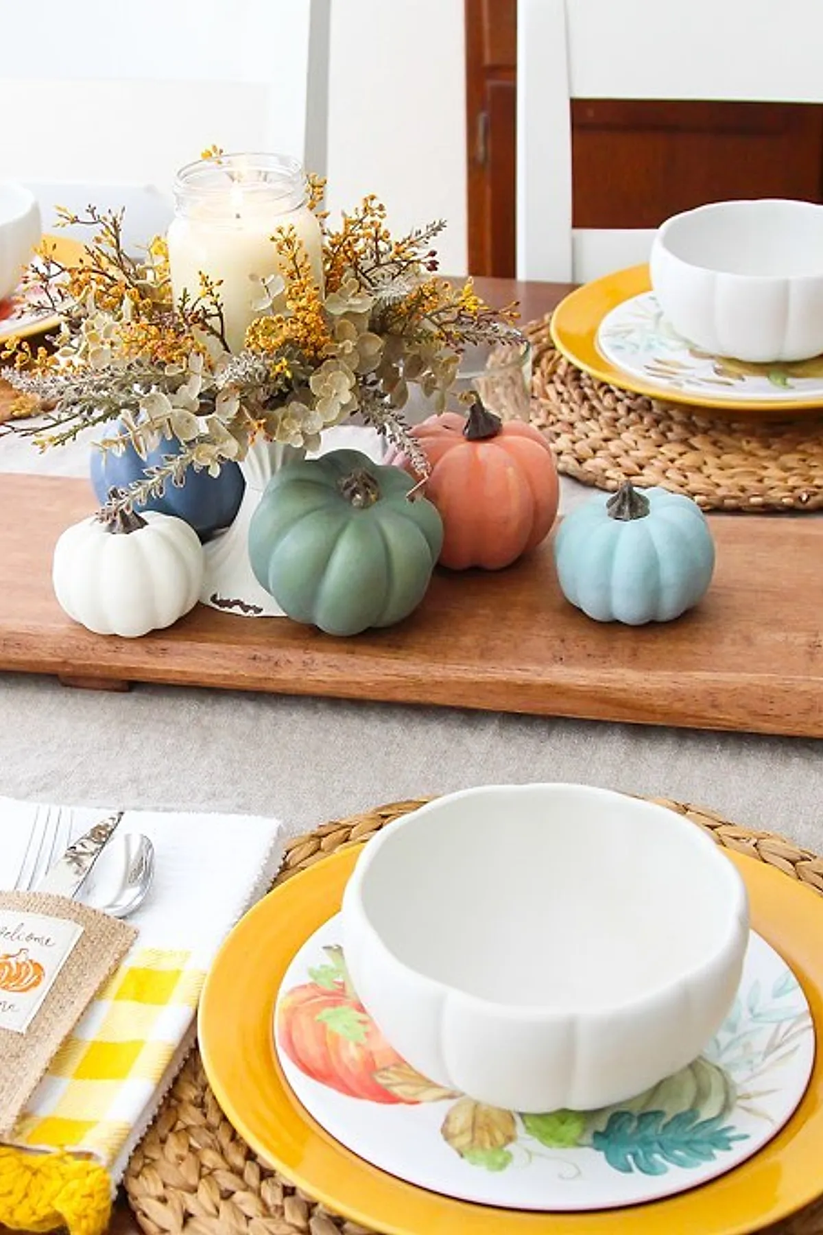 Fall table centerpiece with pastel colored pumpkins in shades of blue, green, peach and white surrounding a glass jar candle with yellow dried flowers in a white pedestal vase; set on a wooden board with yellow rimmed plates and woven placemats.