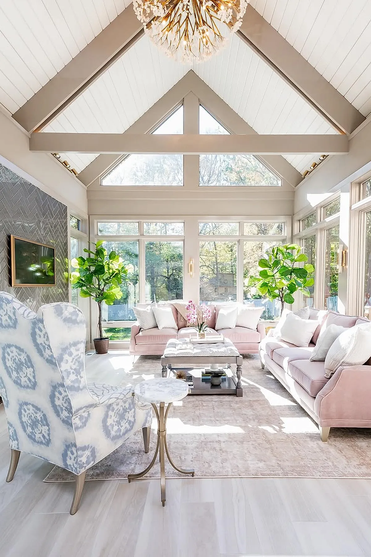 Bright sunroom featuring two pastel pink sofas with white pillows, a blue and white patterned wingback chair, light wood flooring, large windows surrounding the room, tall green plants, and a decorative gold chandelier hanging from a vaulted white ceiling.
