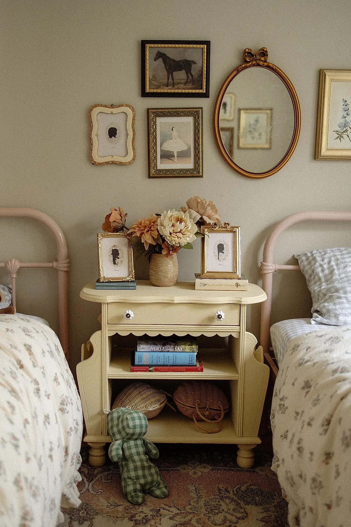 vintage yellow wooden bedside table between two pink metal bed frames, topped with framed floral silhouettes and dried flower arrangement, open shelves holding books and woven baskets, green gingham stuffed doll leaning against table on floral rug