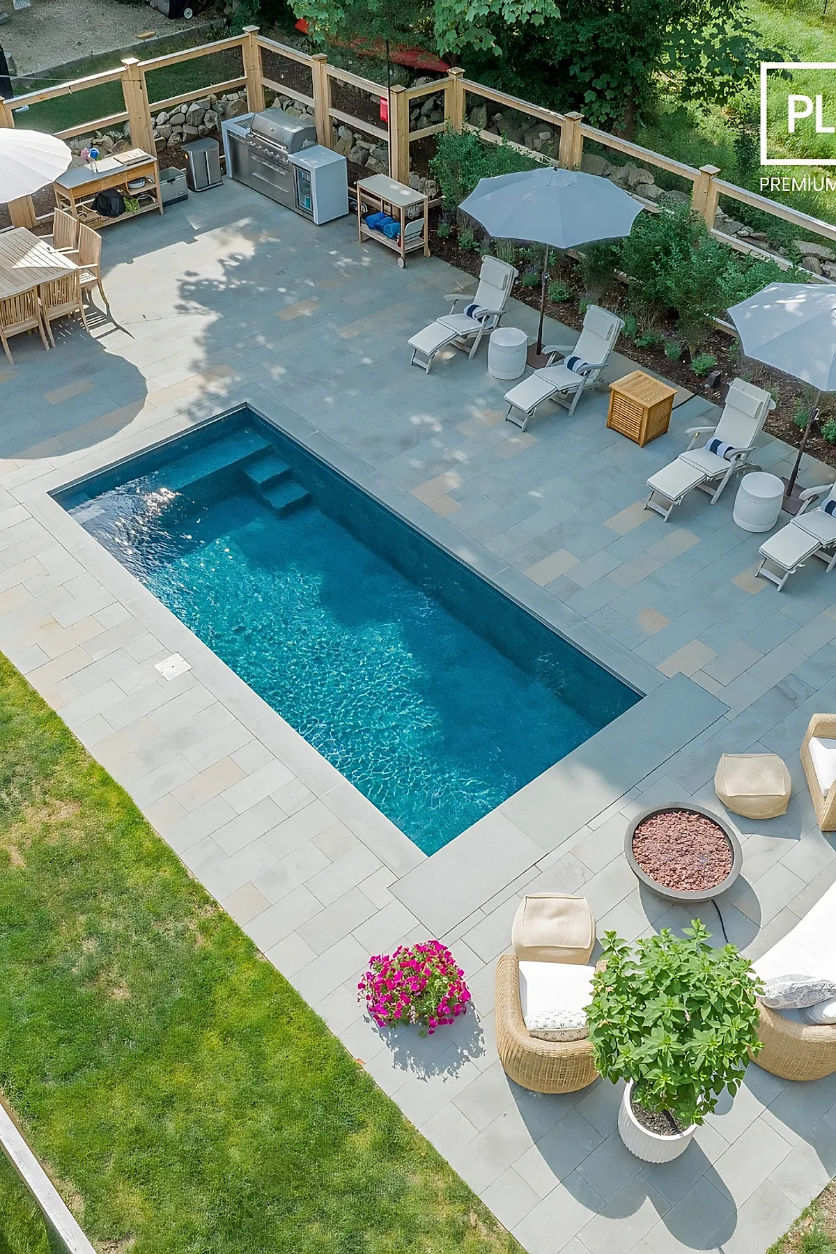 Rectangular modern plunge pool with clear blue water surrounded by light gray stone patio featuring three white cushioned loungers with navy-striped pillows and matching gray umbrellas, adjacent to an outdoor kitchen and dining area enclosed by wooden fencing and lush greenery.