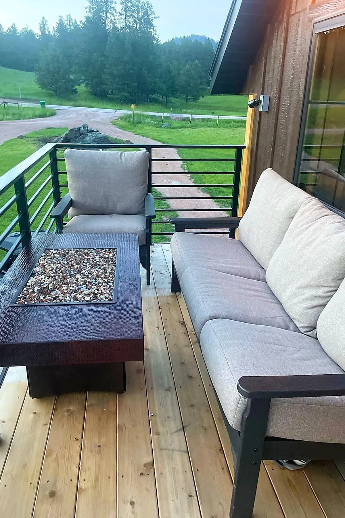 Small outdoor patio with light gray cushioned sofa and chair set around a dark brown fire pit table filled with decorative stones, wooden deck floor, black metal railing, and green grassy landscape in the background.