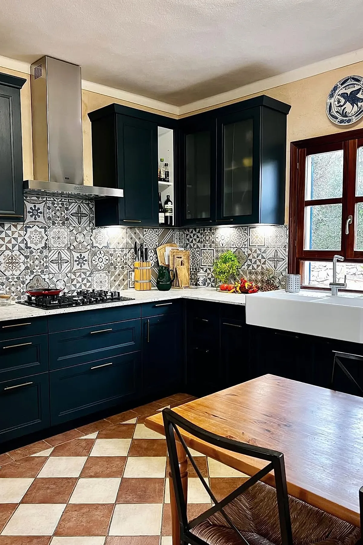 Spanish style kitchen with matte black cabinetry, intricate gray and white patterned tile backsplash, stainless steel range hood, white marble countertops, wooden window frame, and checkerboard terracotta and cream floor tiles.
