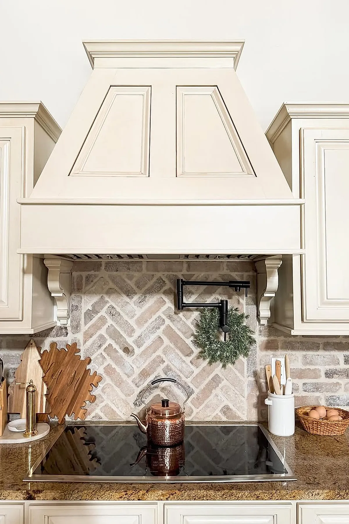 Kitchen featuring a neutral-toned herringbone brick backsplash behind a stovetop with a copper kettle, cream-colored cabinetry, granite countertops, wooden cutting boards, and a small decorative wreath on the wall-mounted black pot filler faucet.
