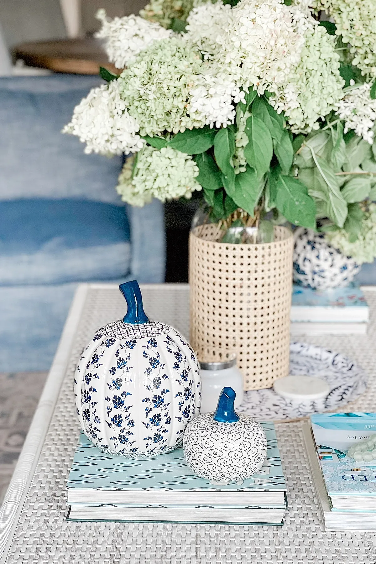 Two ceramic pumpkins with blue floral and geometric patterns on their surfaces, blue stems, sitting atop light blue patterned books; large bouquet of white hydrangeas in woven vase behind them, set on a woven textured table surface.