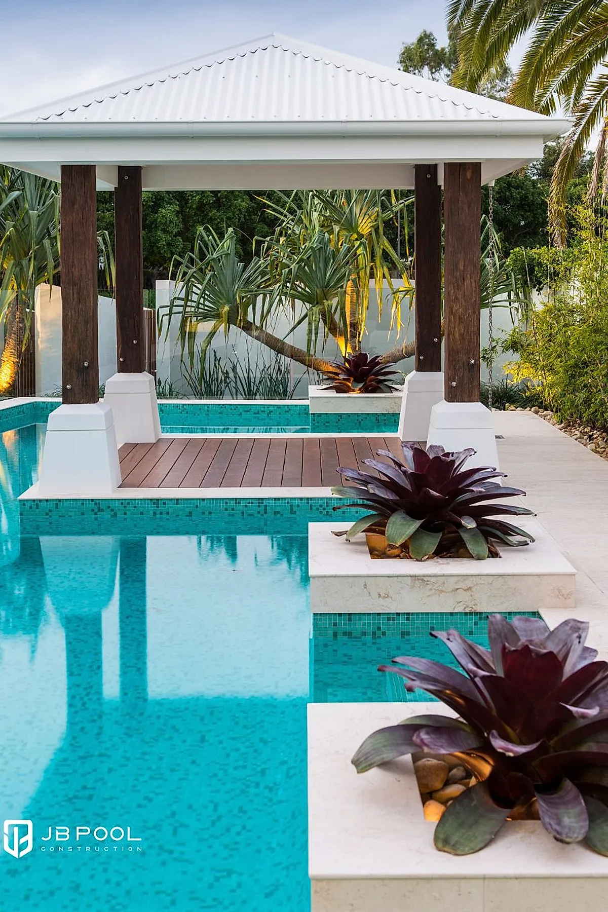 Modern pool deck featuring a wooden pavilion with dark wood pillars and a white roof, surrounded by turquoise mosaic tile pool water and square planters holding large purple-green succulent plants, with palm trees and lush greenery in the background.