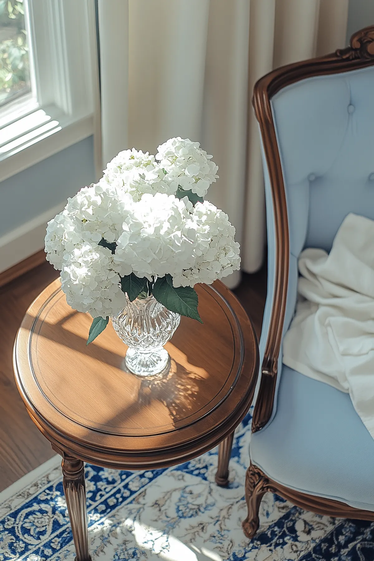 A crystal vase filled with white hydrangeas on a wooden side table next to a blue armchair.