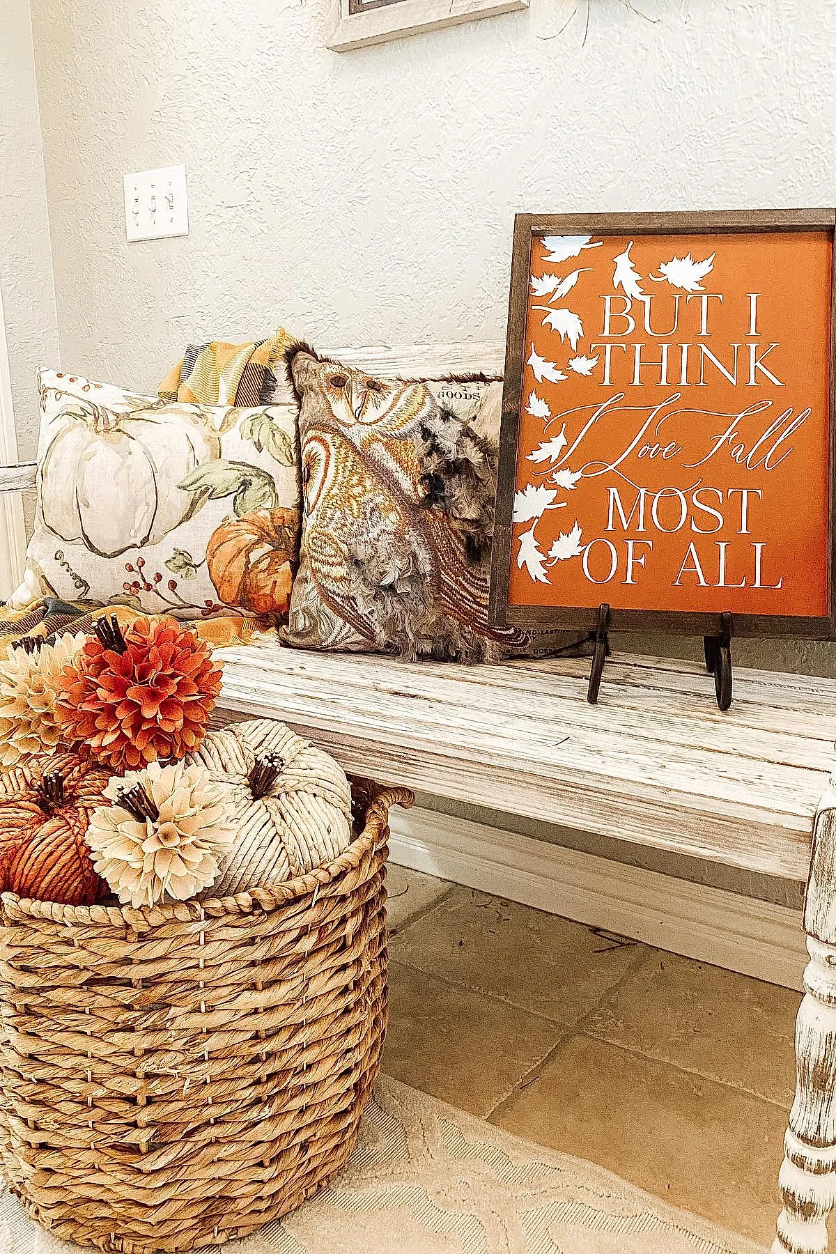 White wooden bench holding layered fall-themed pillows including a floral pumpkin print pillow and a feather-textured pillow next to an orange framed sign reading