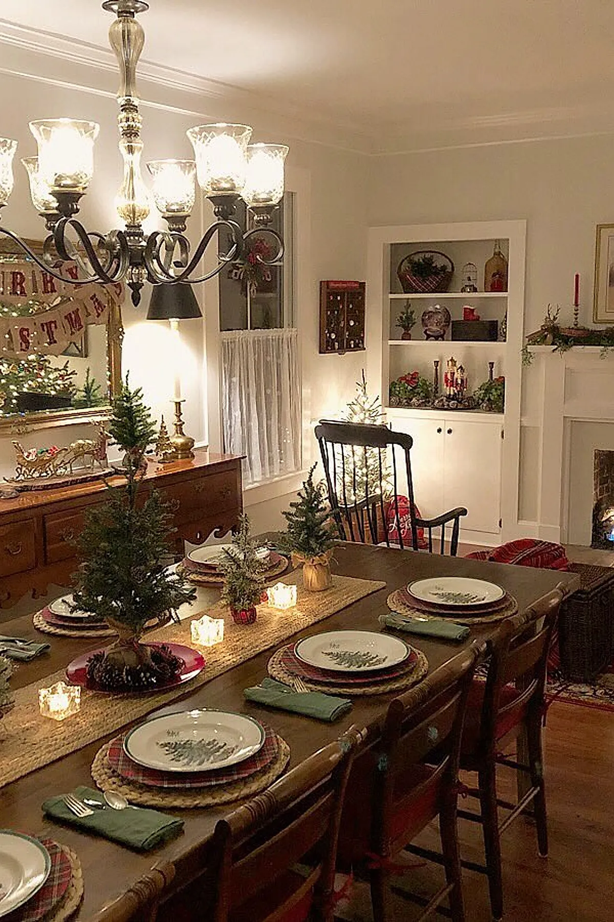 Rustic wooden Christmas dining table with small potted pine trees, plaid placemats, white plates with tree designs, green napkins, and glowing votive candles, surrounded by warm holiday decor and a lit Christmas tree.