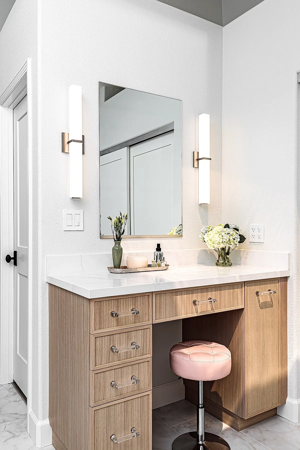 wooden bedroom vanity with white marble top, square frameless mirror flanked by two vertical white wall sconces, pink tufted round stool with chrome base, green vase with flowers, glass bowl with white flowers