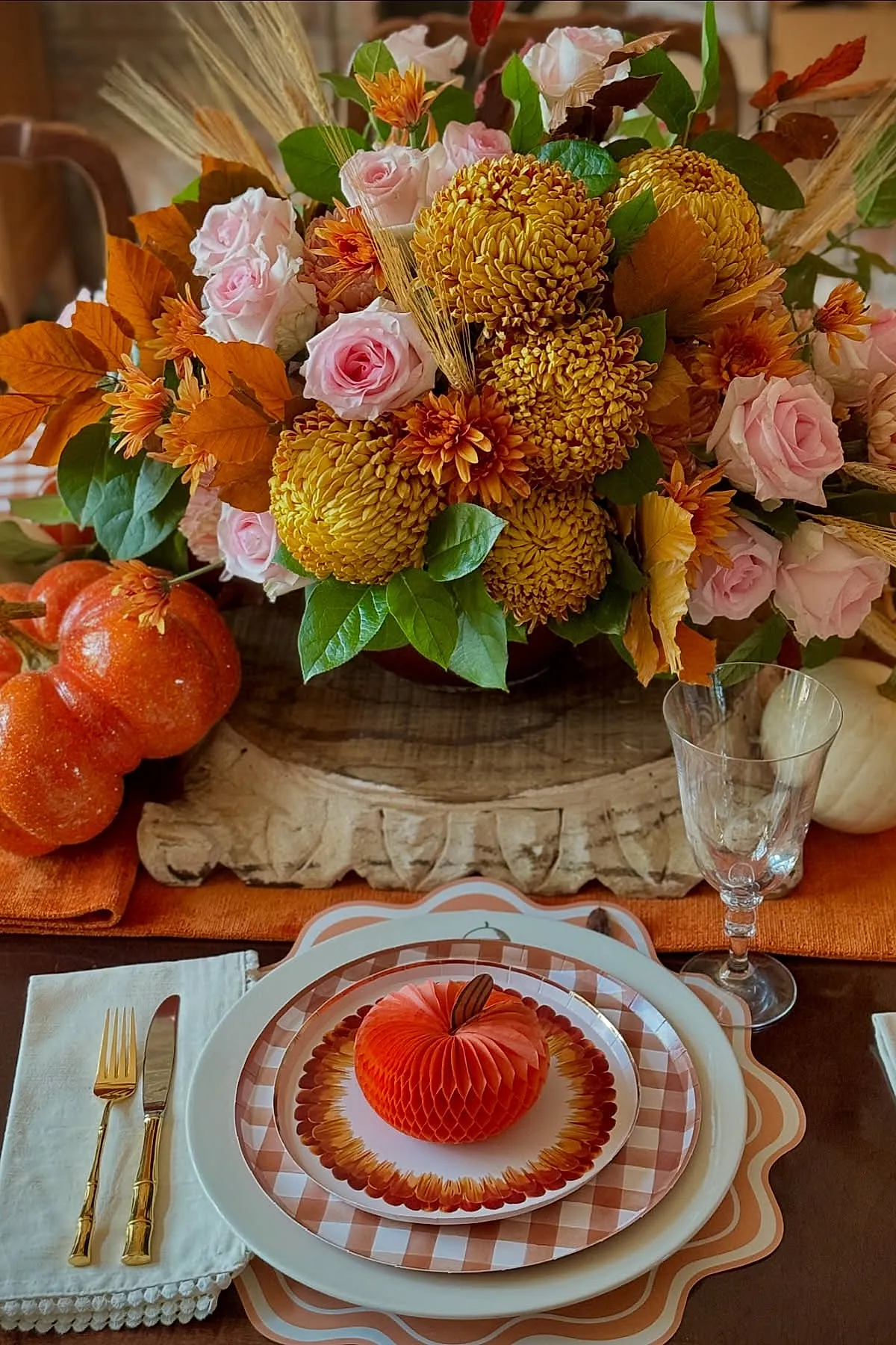 Thanksgiving table centerpiece featuring pink roses, golden chrysanthemums, orange leaves, green foliage, wheat stalks, clear wine glass, paper pumpkin on patterned plates with gold flatware on white napkin