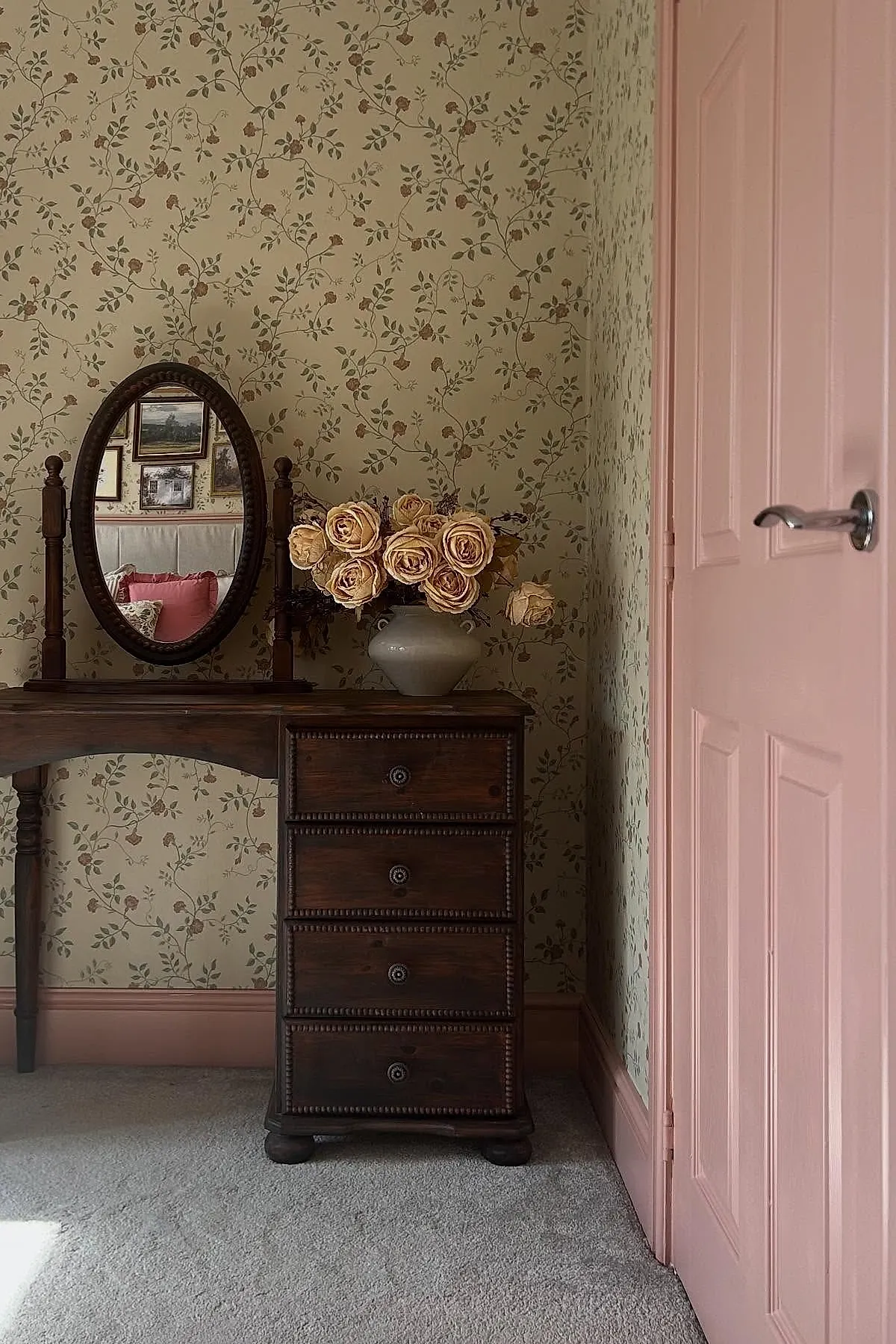 dark wooden vanity with oval mirror next to pale pink door, floral wallpaper in soft green and brown tones, beige vase holding muted pink dried roses on vanity