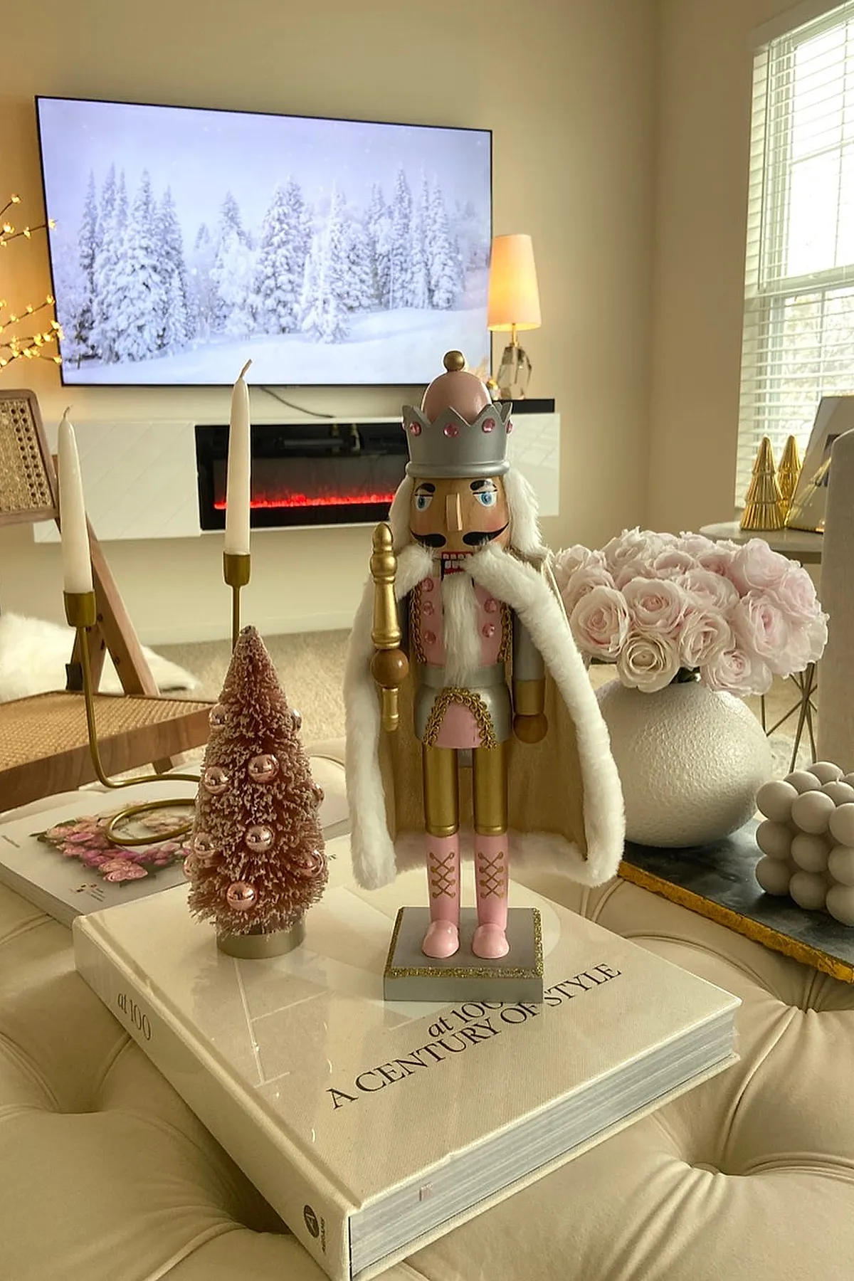 christmas coffee table decor featuring a pink and gold nutcracker figure, a small pink bottle brush Christmas tree with rose gold ornaments, and a white textured vase filled with pale pink roses on top of a large hardcover book on an upholstered ottoman.