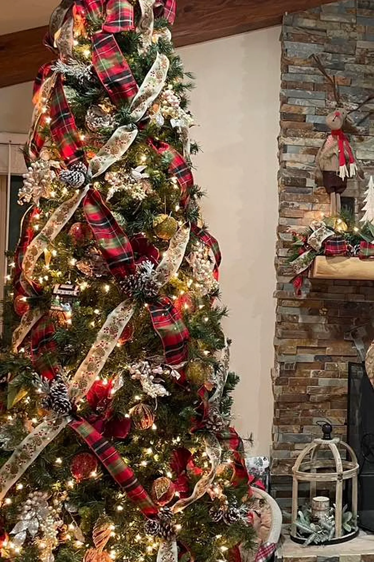 A Ralph Lauren Christmas tree decorated with wide red plaid and cream ribbons, frosted pinecones, gold ornaments, white berries, and warm white lights standing next to a stone fireplace with rustic holiday decor including a reindeer figure and greenery accented with matching plaid ribbon.