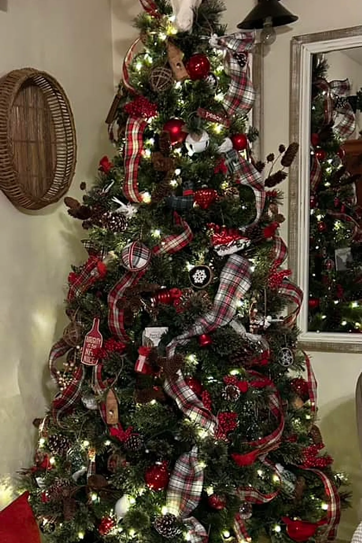 A Ralph Lauren Christmas tree decorated with red and cream plaid ribbons, pinecones, red berries, ornaments, and white lights near a neutral wall with a woven wall basket.