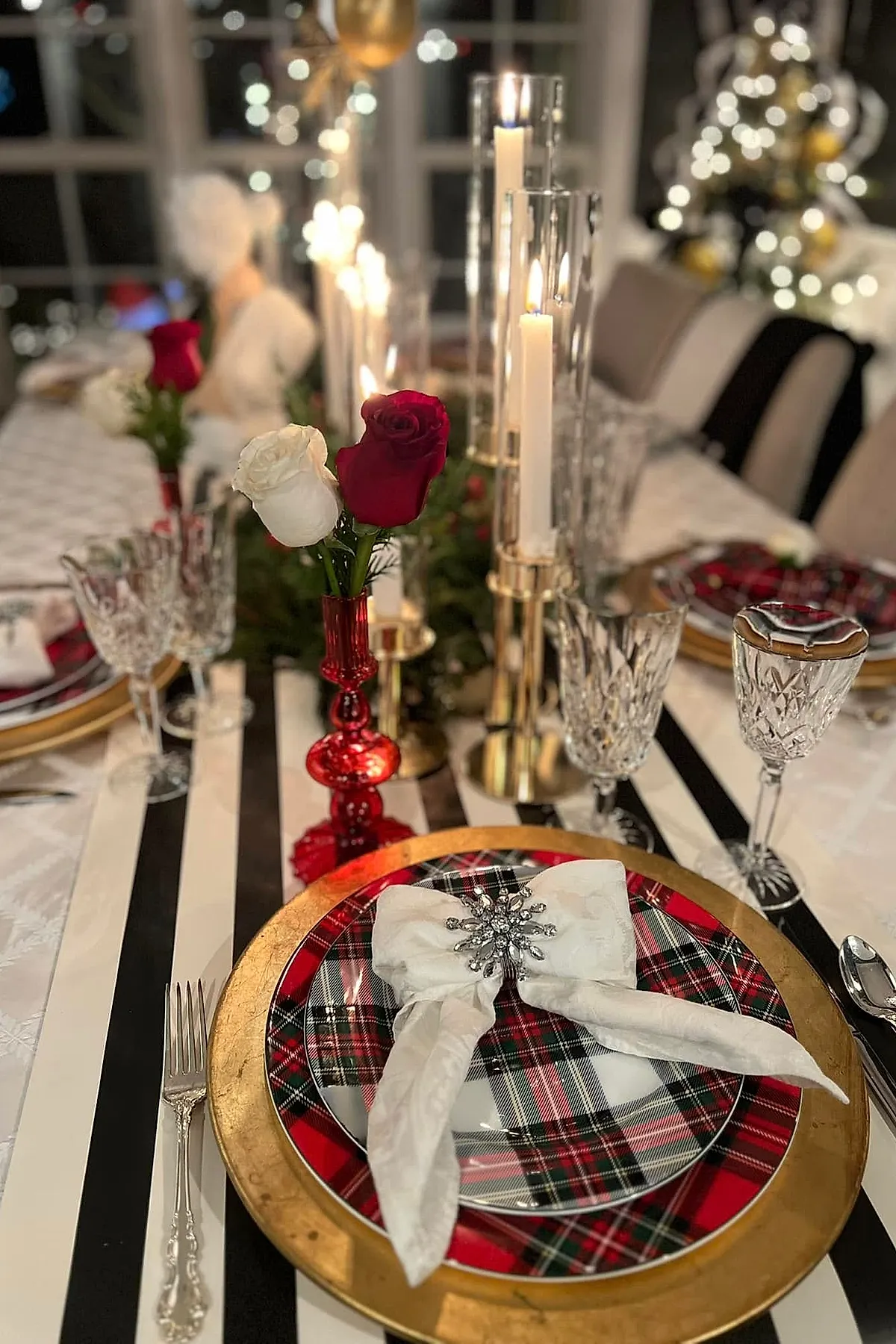 A Ralph Lauren Christmas table setting featuring red and white plaid plates on a gold charger, white napkin with jeweled brooch, crystal glassware, red and white roses, gold candle holders, and striped runner.