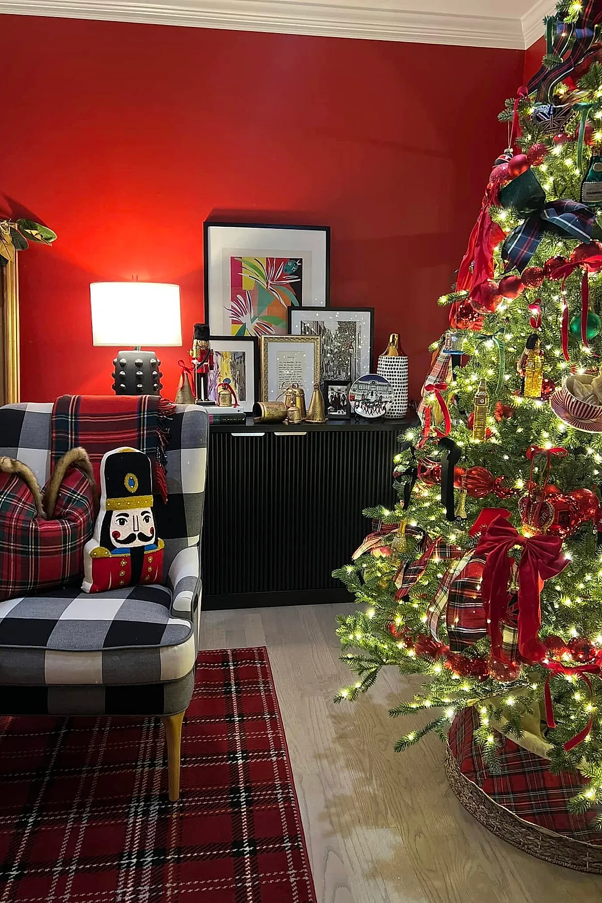 A Ralph Lauren Christmas tree decorated with plaid ribbons, red bows, and ornaments beside a black and white checkered chair with a nutcracker pillow and plaid throw against a red wall and black cabinet with framed art.