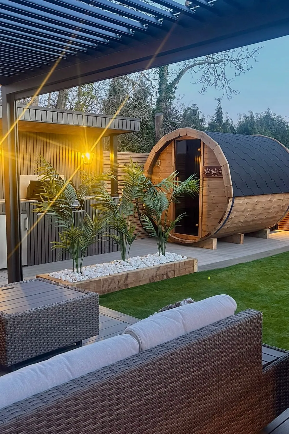 Outdoor patio with wooden planter privacy wall containing green plants and white stones, adjacent to a wooden barrel sauna under a pergola with warm lighting