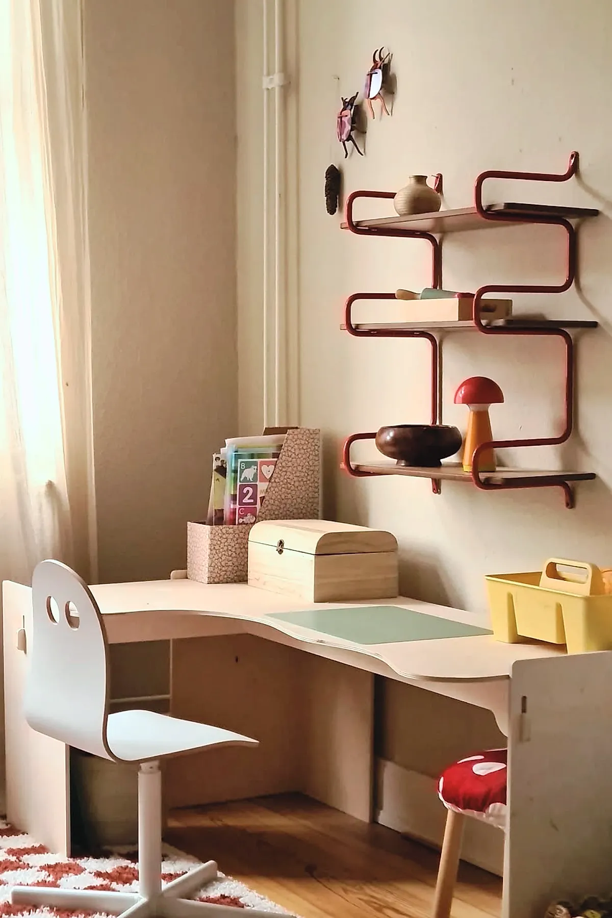 Kids desk area in bedroom featuring a curved wooden kids study desk with light wood finish, white swivel chair with round cutouts on the backrest, wall-mounted red metal shelving holding decorative objects including a wooden bowl and mushroom lamp, soft natural light coming through sheer curtains.