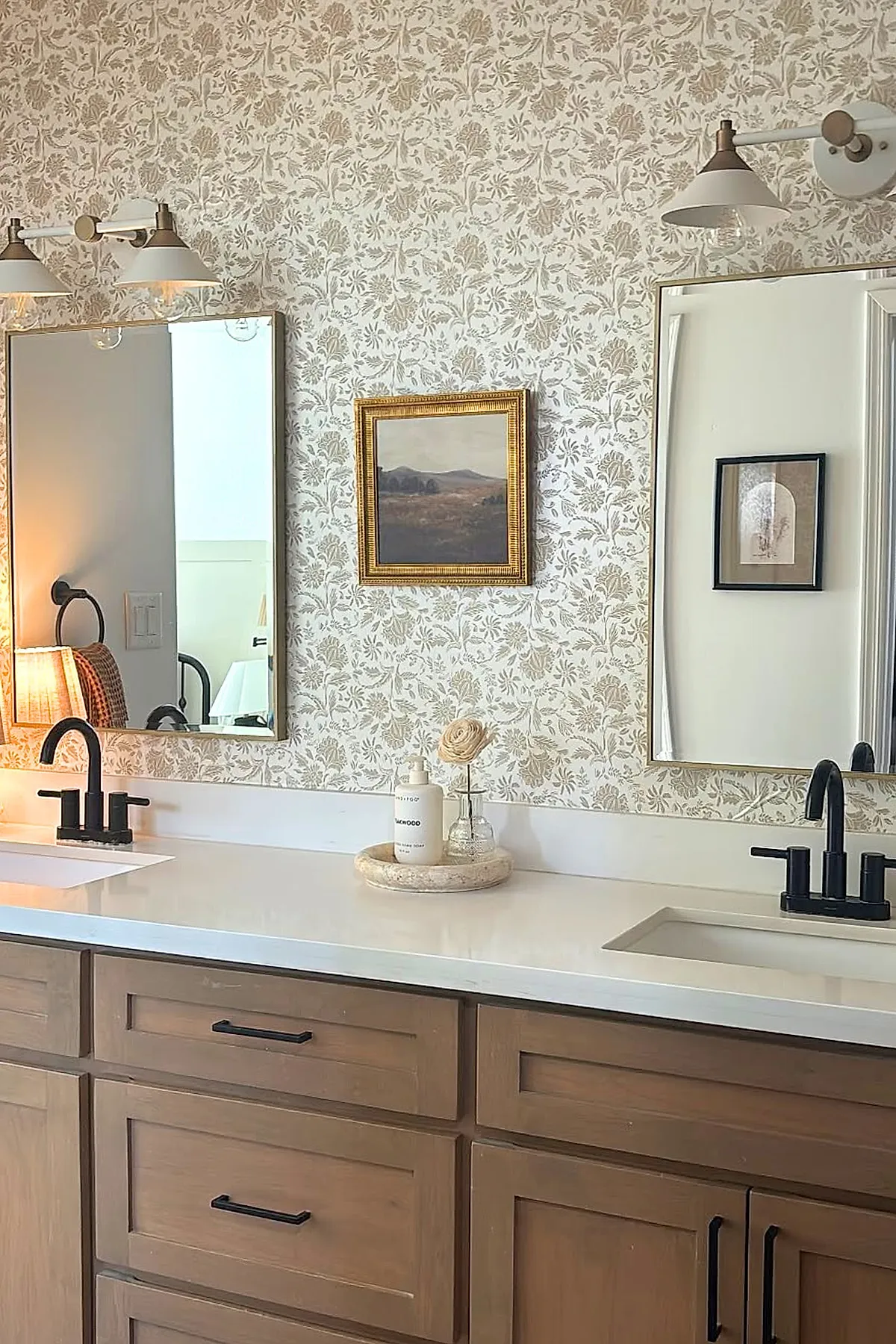 Warm neutral bathroom featuring floral patterned wallpaper in soft beige and cream tones above a white countertop with dual sinks, wood vanity cabinets with black handles, two rectangular mirrors framed in gold, and modern light fixtures above each mirror.