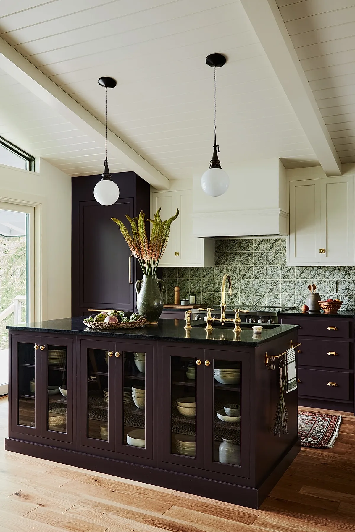 Cottage kitchen featuring dark purple lower cabinets and island with glass front doors, white upper cabinets, green patterned vintage tile backsplash behind stove, brass faucet and hardware, two globe pendant lights hanging from a white paneled ceiling, wooden floor, and a vase with tall flowers on the island countertop.
