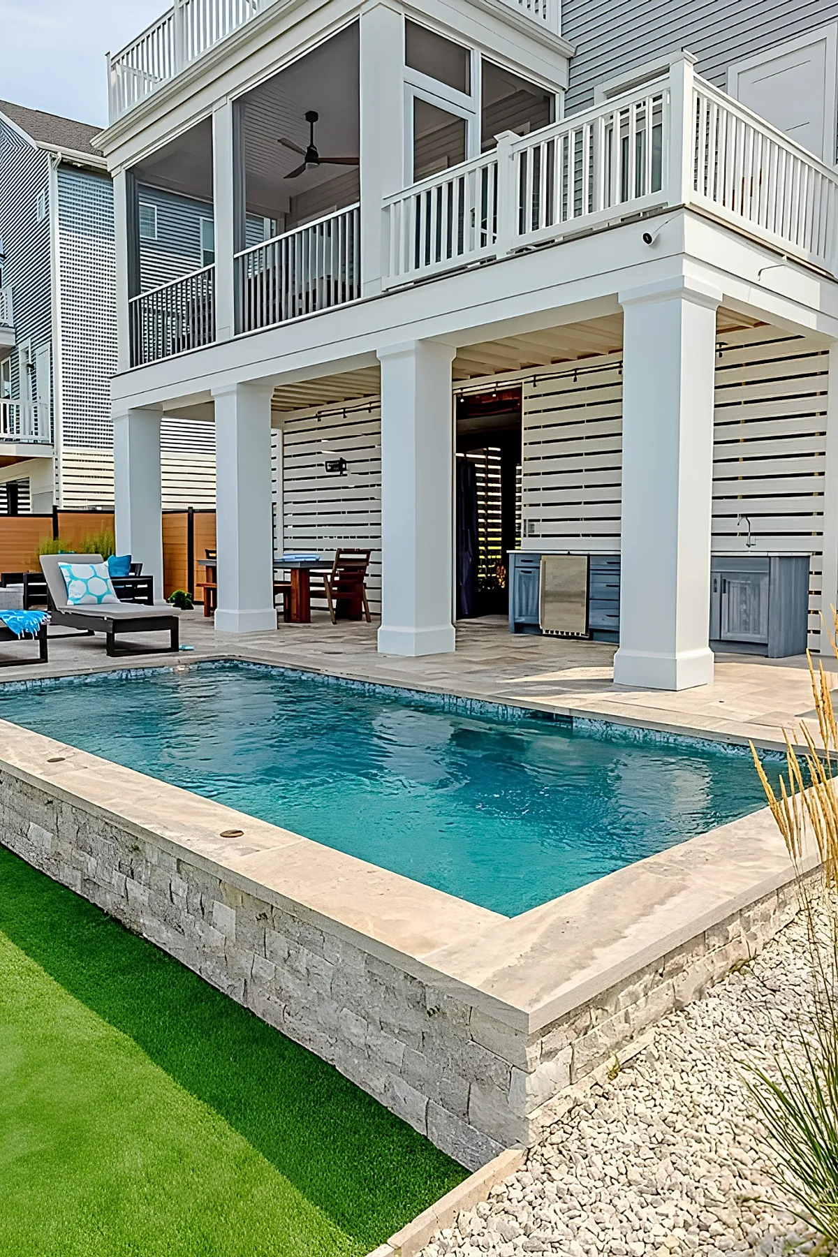Modern rectangular plunge pool with clear blue water surrounded by stone coping and light beige tiled patio beneath a white covered deck supported by large square columns, featuring outdoor furniture and an outdoor kitchen area in the background.