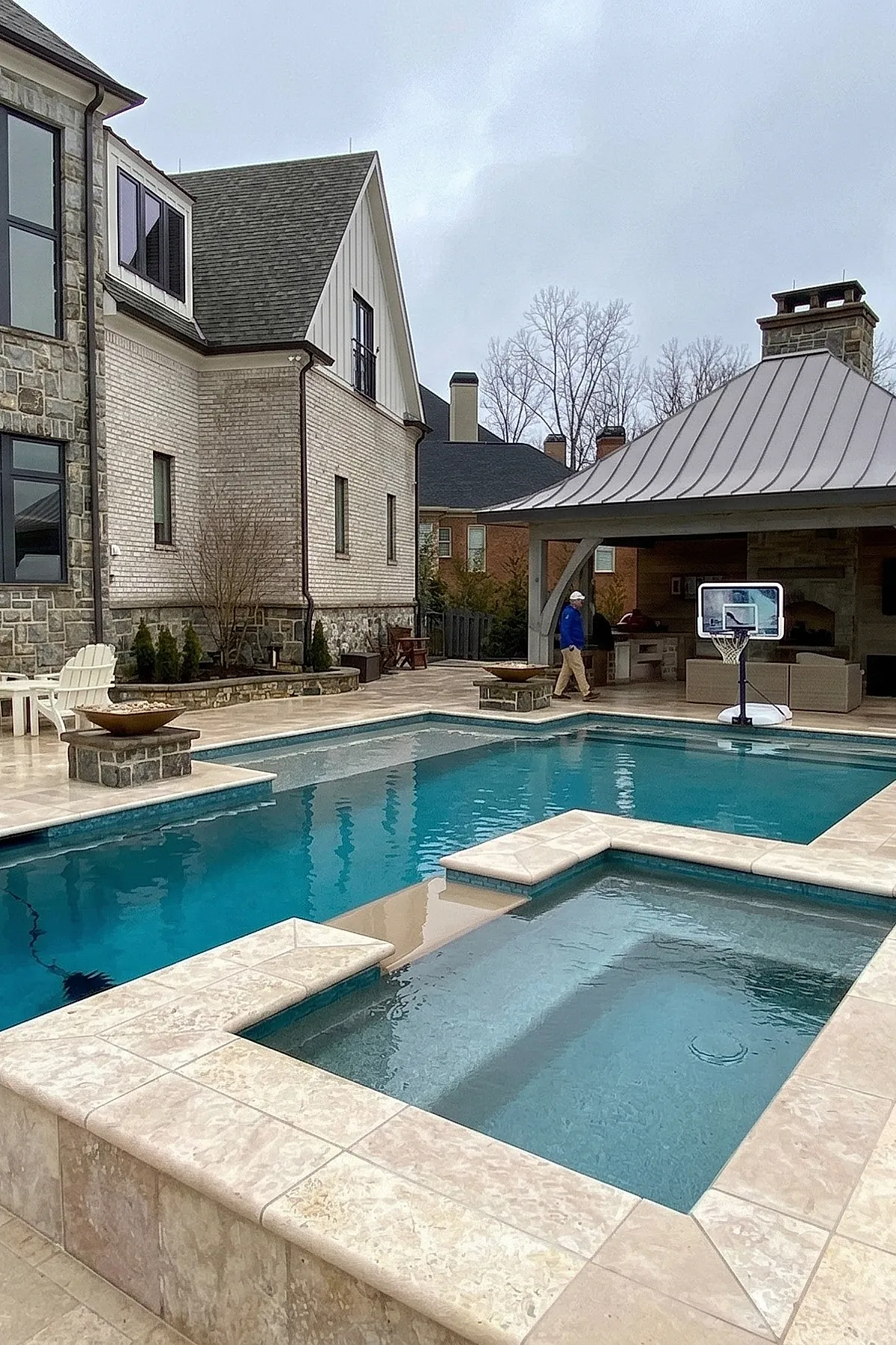 Elegant pool deck featuring a rectangular swimming pool with an attached hot tub, surrounded by light beige stone tiles, next to a covered lounge area with seating and a basketball hoop in the background under overcast sky.