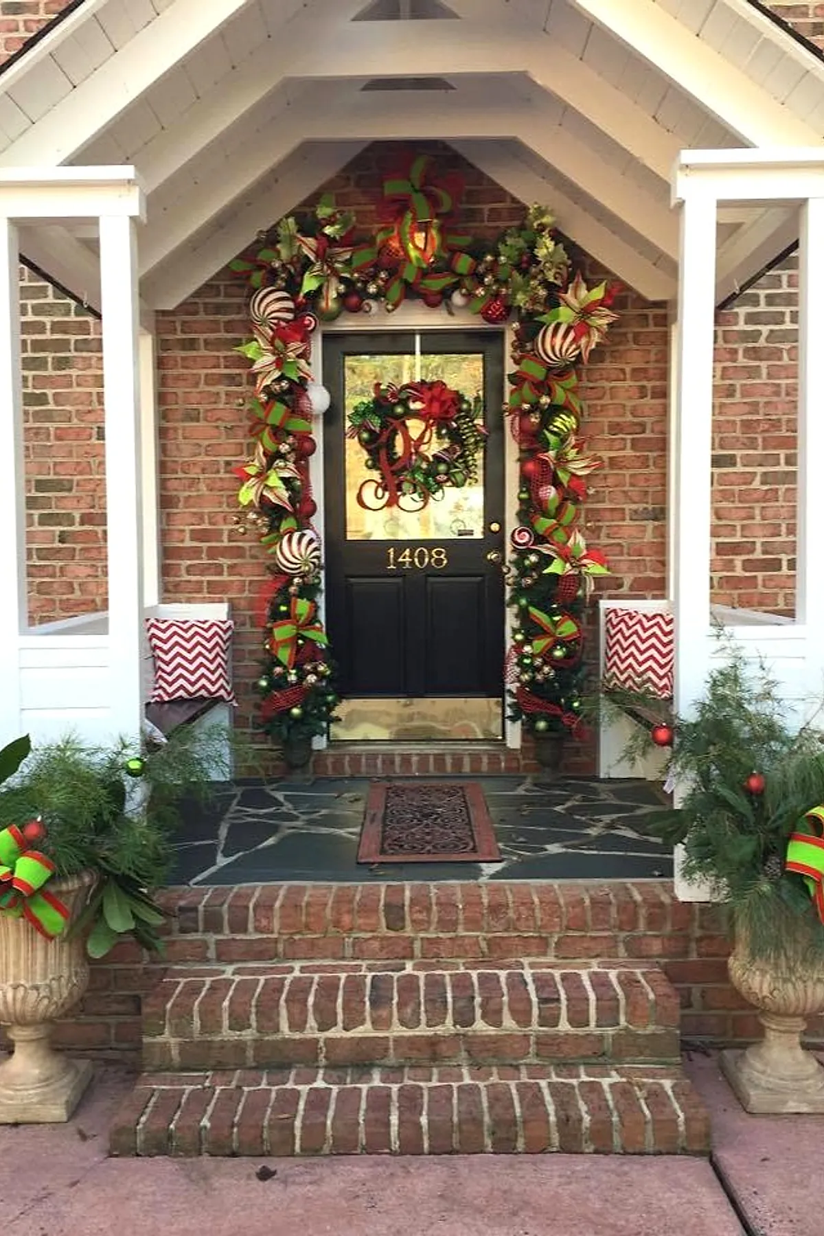 Front porch decorated with a thick garland around the door frame featuring large red and green striped ribbons, big ornaments in red, green, gold, and white, paired with matching wreath on black door; greenery-filled stone urns with similar ribbon accents flank brick steps.