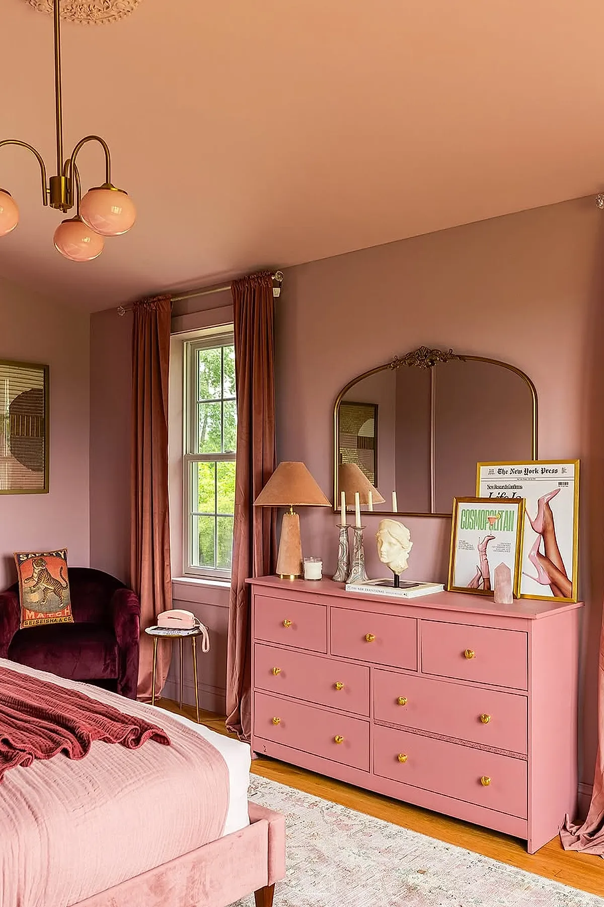Warm pink bedroom featuring a dusty rose dresser with gold knobs, matching pink curtains, a velvet burgundy chair, soft pink bed linens, and decorative art pieces on the dresser under a brass-framed mirror.