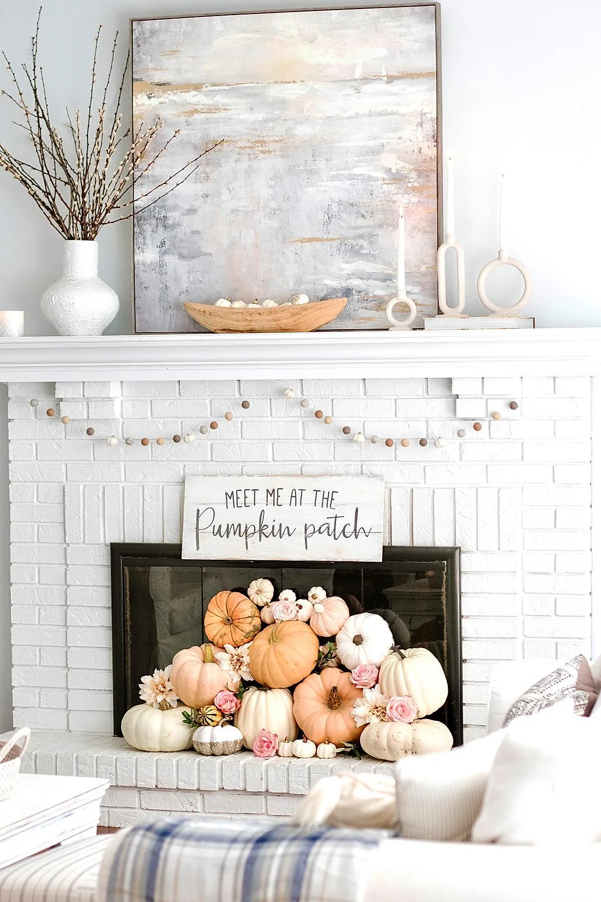 white brick fireplace with neutral-toned pumpkins and flowers stacked inside the firebox under a wooden sign that says meet me at the pumpkin patch, surrounded by minimal white decor on the mantel including abstract candlesticks and a textured vase with branches
