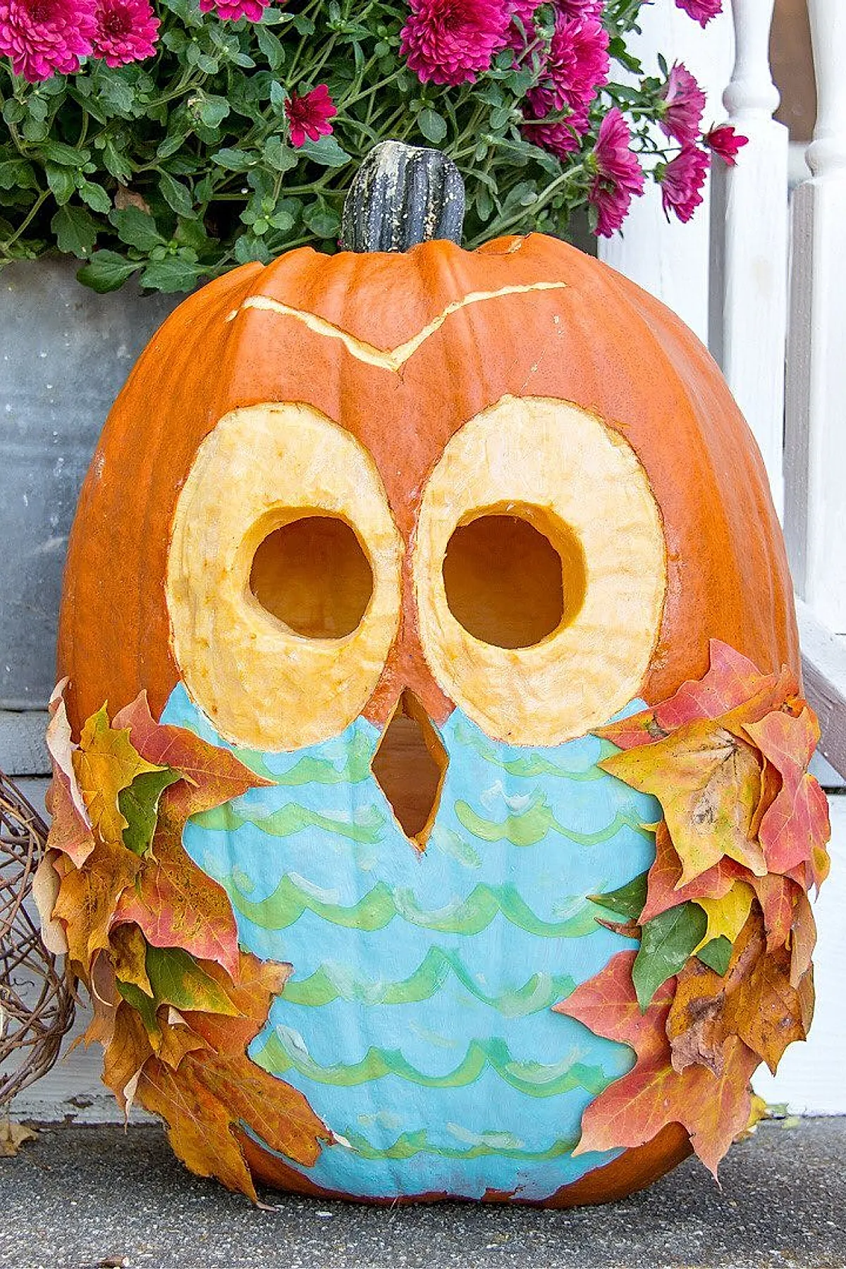 Orange pumpkin carved with large round eyes and beak, painted blue and green owl body, decorated with autumn leaves attached as wings, set against pink flowers.