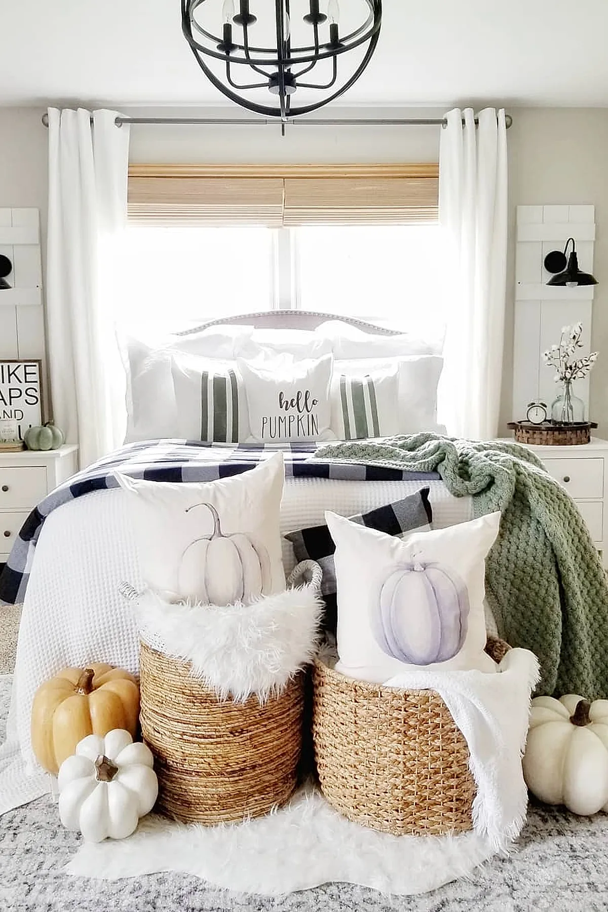 bedroom with white bedding and green throw, plaid blanket, two woven baskets holding white pillows with pumpkin illustrations, assorted cream and beige pumpkins on white rug