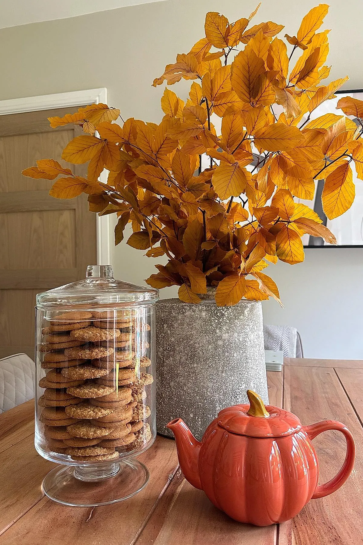 Wooden table featuring a tall gray stone vase filled with bright orange autumn leaves, next to a tall clear glass jar filled with stacked cookies, and an orange ceramic teapot shaped like a pumpkin.