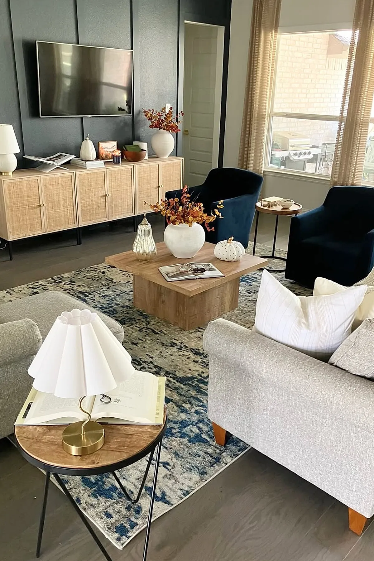 Earthy living room featuring a light wood cabinet with woven doors, two dark navy velvet armchairs, beige sofa with white pillows, wooden coffee table decorated with white ceramic pumpkins and a vase with orange foliage. The room has dark accent walls behind the cabinet and large windows with beige curtains letting in natural light.