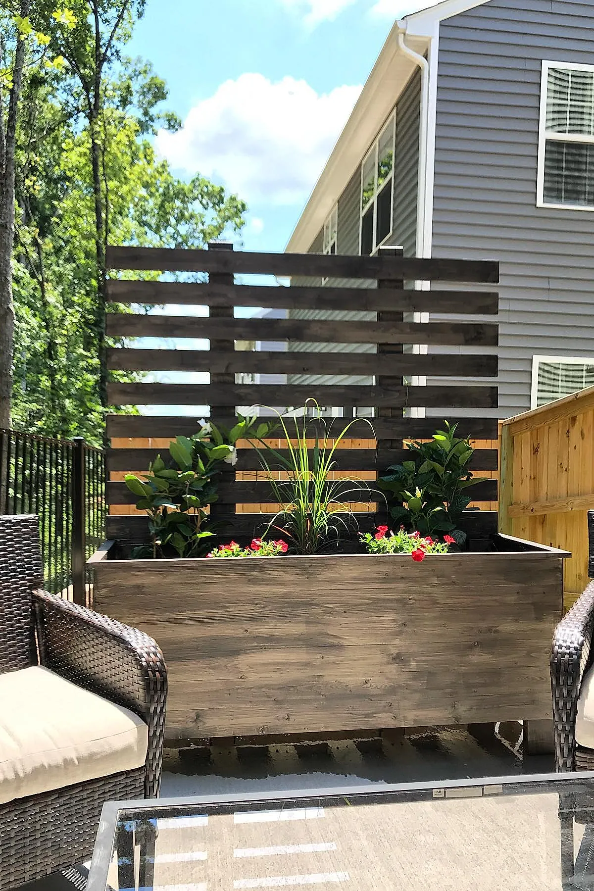 Wooden planter privacy wall with horizontal slats containing green plants, set on an outdoor patio next to wicker chairs