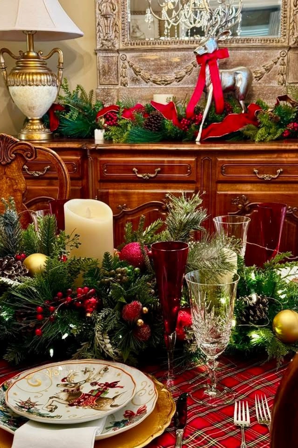 A Ralph Lauren Christmas dining table decorated with a red plaid tablecloth, gold chargers, reindeer plates, crystal glassware, and lush greenery with red and gold ornaments and candles. In the background, a wooden sideboard is adorned with garland, pinecones, and a silver reindeer statue with a red bow beneath an ornate mirror.
