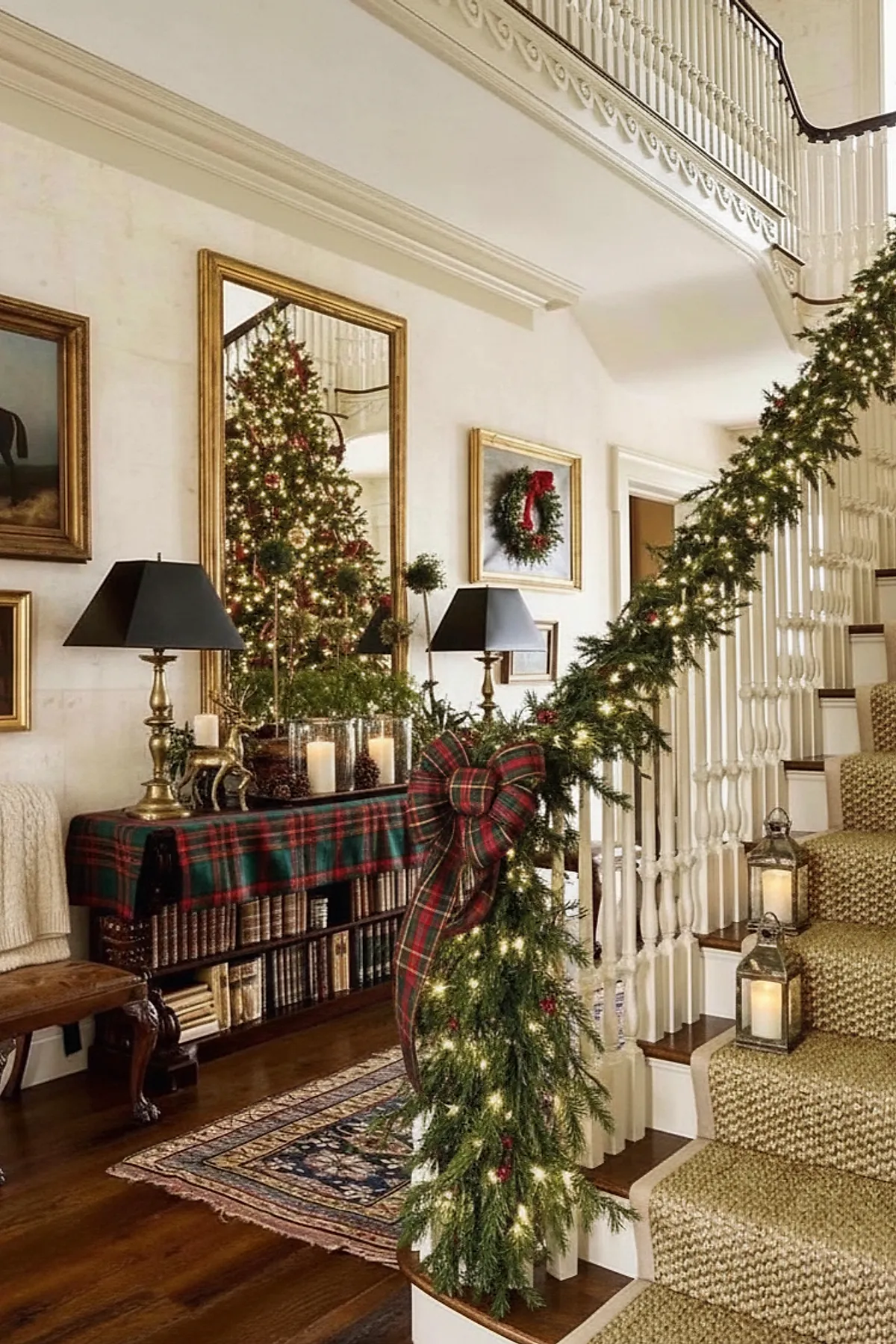 A Ralph Lauren Christmas staircase featuring lush green garland wrapped around white banisters with warm white lights and a large red and green plaid bow, brass lanterns with candles, and a console table draped in matching plaid fabric with candles and greenery beneath a gold-framed mirror reflecting the decorated tree.
