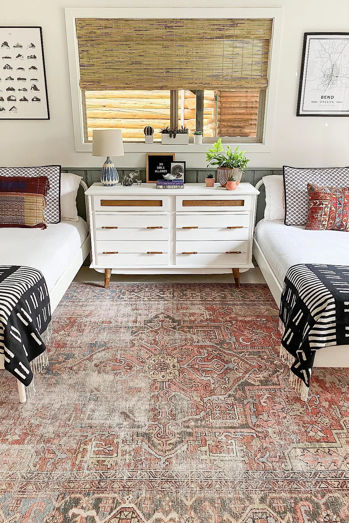Two twin beds with white bedding featuring layered patterned pillows in red, blue, and beige flank a white dresser topped with potted plants, lamp, and books. Natural bamboo blinds cover the window above, framed by light-colored walls with black framed artwork on each side. A faded terracotta patterned rug covers the floor.