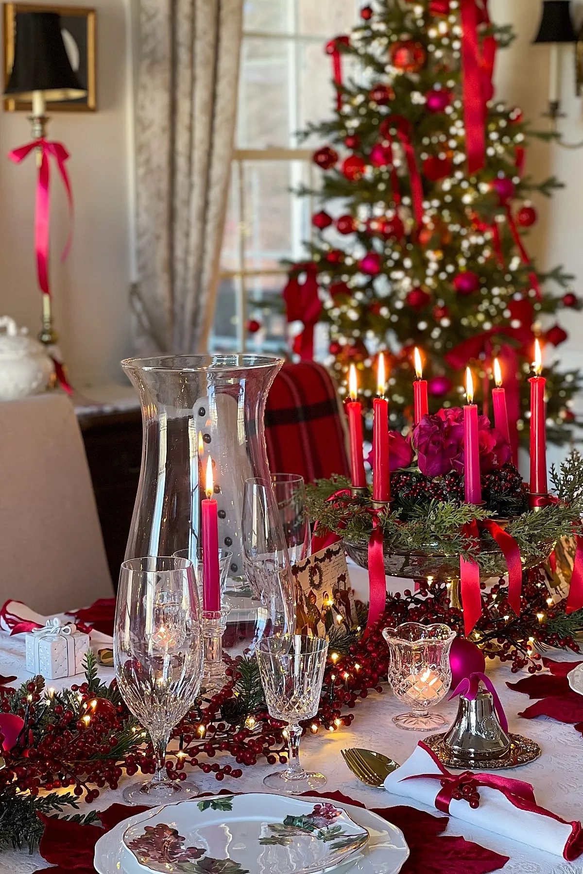 christmas dinner table settings featuring red taper candles in a wreath centerpiece, red berries garland, crystal glassware, white plates with holly pattern, gold flatware, white napkins tied with red ribbon and berries, set against a backdrop of a decorated Christmas tree with red ornaments and lights.
