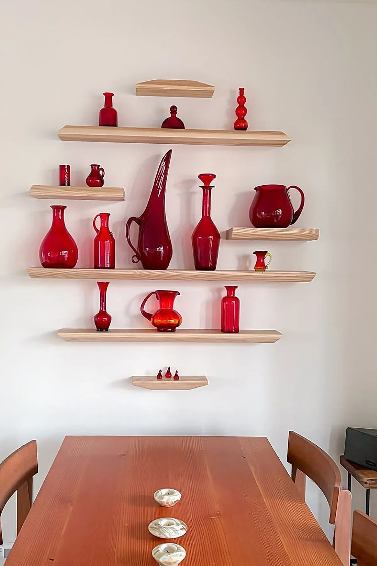 Light wood floating shelves arranged in a geometric pattern on a white wall displaying an array of red glass vases, pitchers, and bottles above a wooden dining table with small decorative bowls.