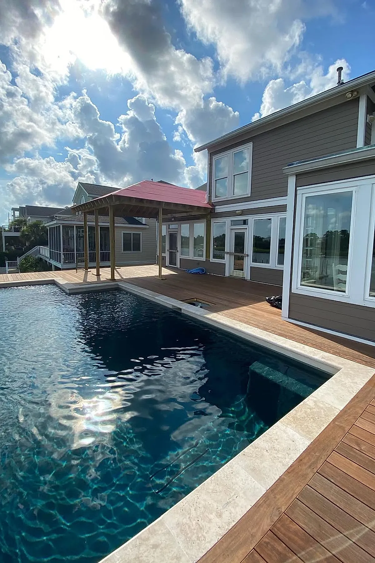 Swimming pool with dark blue water bordered by a light stone edge and surrounded by a wooden pool deck featuring a covered seating area with a red roof attached to a two-story gray house with multiple windows.