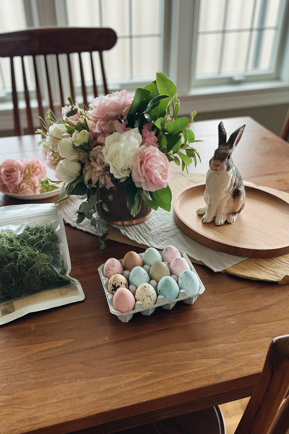 Elegant dining table with roses, a ceramic rabbit, speckled eggs, and natural light from large windows.