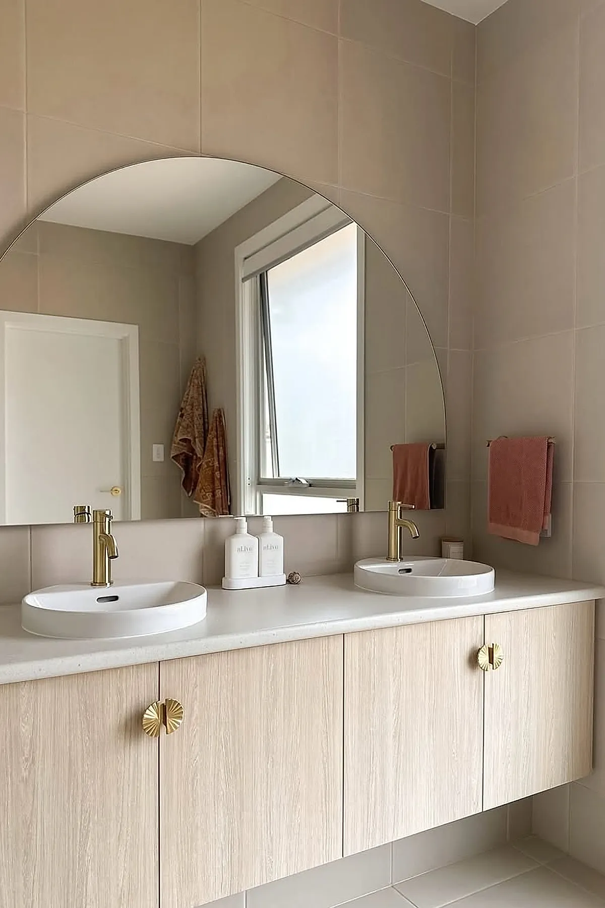 Large round bathroom mirror mounted above a floating light wood vanity with dual white vessel sinks and brushed brass faucets, set against neutral beige tiled walls with matching gold cabinet knobs.