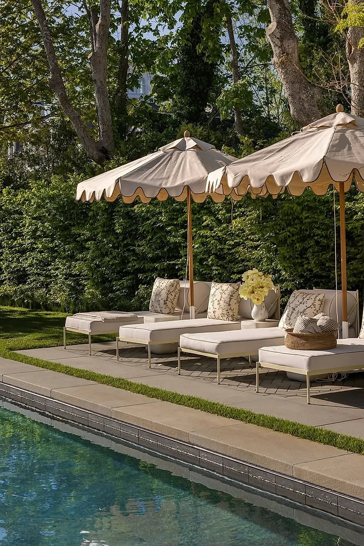 Pool deck featuring beige cushioned lounge chairs with patterned pillows under cream-colored scalloped-edge umbrellas, surrounded by lush green trees and hedges next to a clear swimming pool.