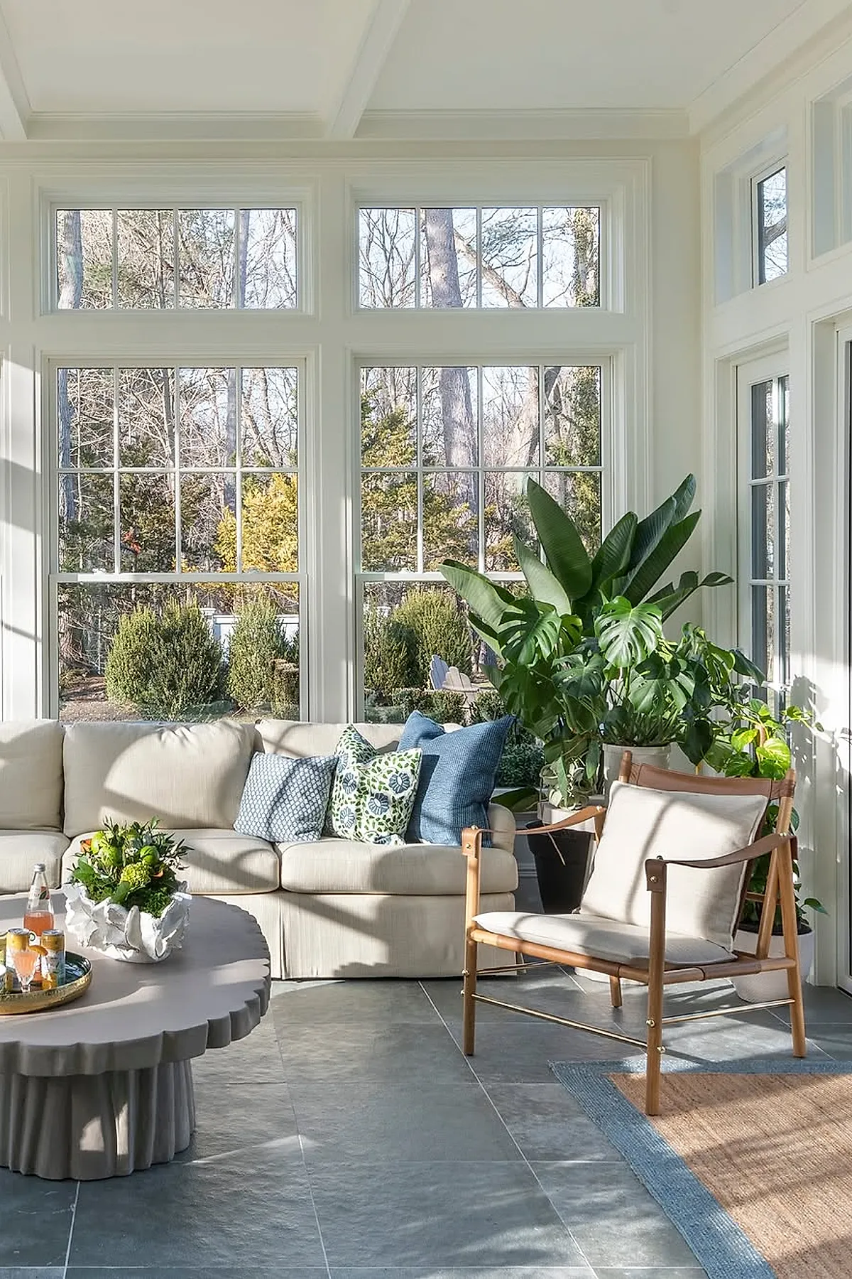 Bright sunroom featuring a light beige sectional sofa with blue and patterned throw pillows, a sculptural round coffee table with a plant centerpiece, wooden framed armchair with cream cushions, large windows showcasing greenery outside, and various large green plants clustered in the corner.