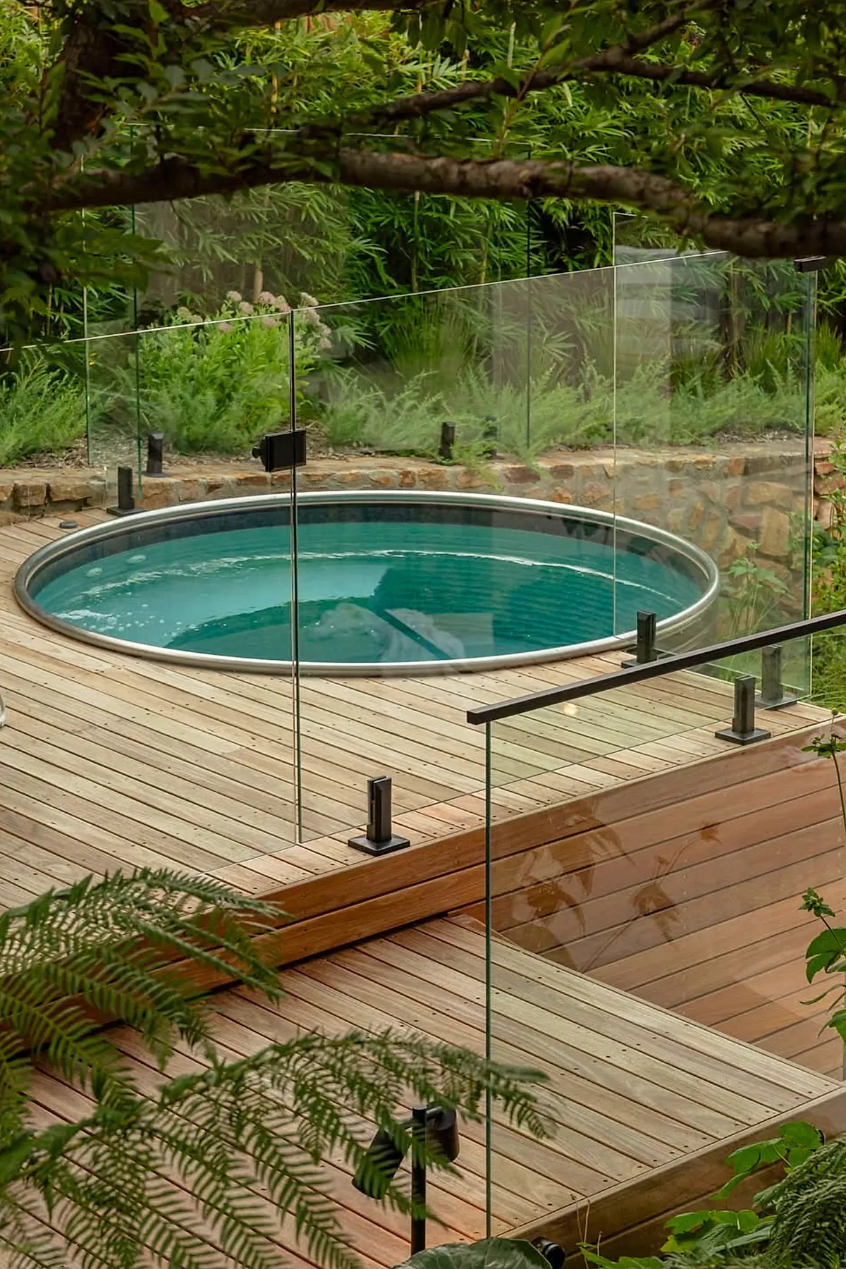 Round above ground plunge pool with blue water surrounded by a wooden deck and clear glass safety fencing, set against lush green plants and stone wall backdrop.