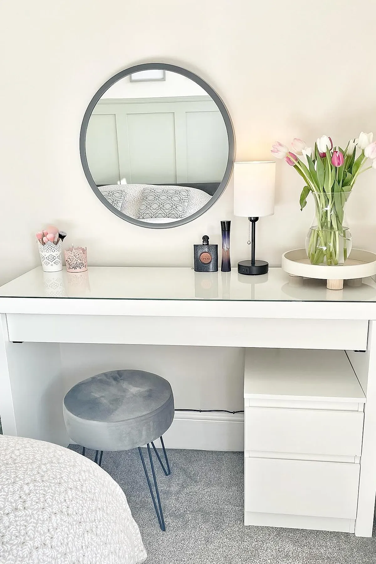 white vanity desk with glass top, round gray-framed mirror on wall, gray velvet stool with metal legs, white lamp with black base, clear vase holding pink and white tulips on tray