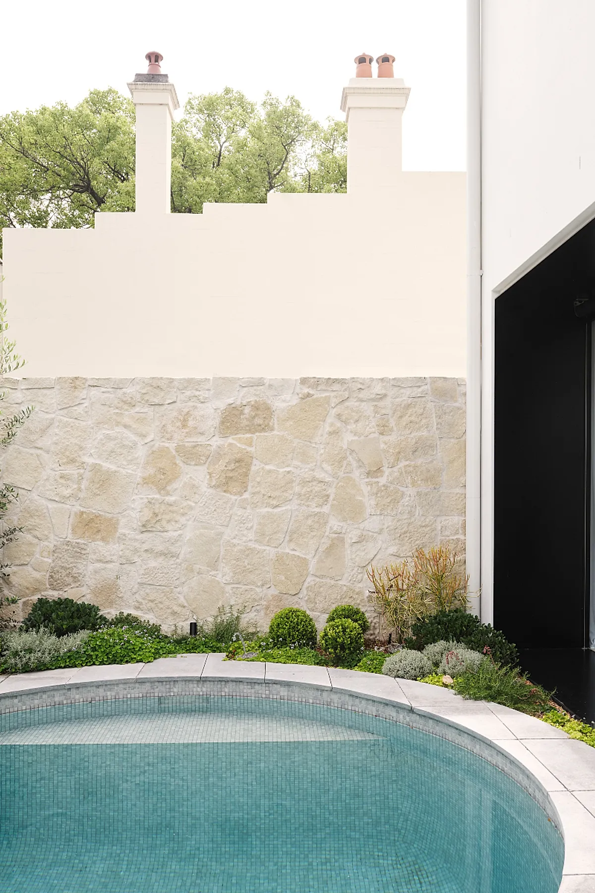 Round plunge pool with blue mosaic tiles surrounded by a curved white stone edge, adjacent to a beige irregular stone wall and lush green garden plants in the background.